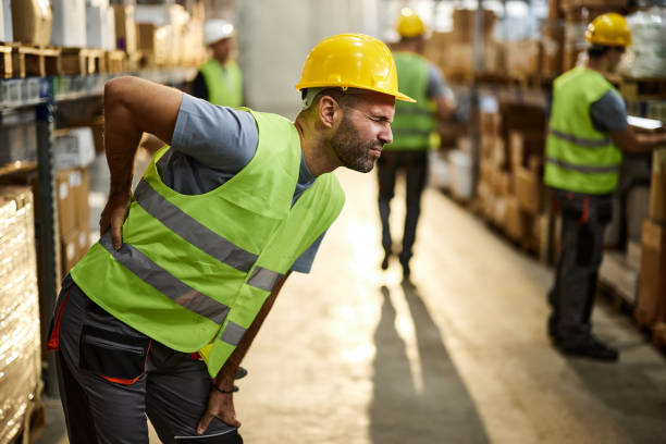 Displeased manual worker holding his back in pain at distribution warehouse. His colleagues are in the background.